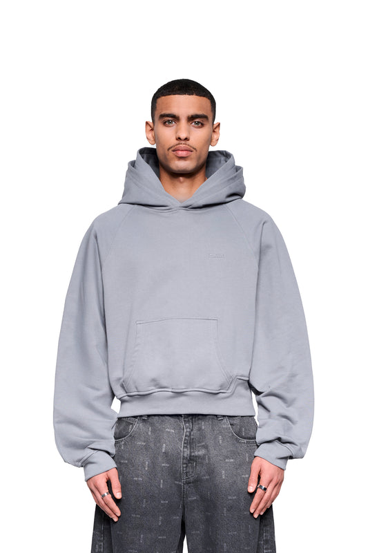 Ein junger Mann trägt den OLAKALA Double Hood Hoodie Gray mit Kängurutasche, Raglan-Ärmeln und asymmetrischem Bund, kombiniert mit locker geschnittenen Jeans.