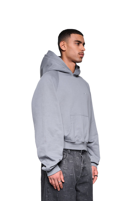 Junger Mann im Profil trägt Double Hood Hoodie Gray mit Kängurutasche und Raglanärmeln, kombiniert mit charcoal-washed Jeans, zeigt schlichte Silberringe an der linken Hand.