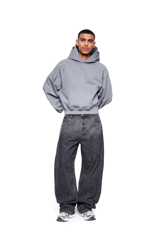 Männliches Model trägt Double Hood Hoodie Gray mit Kängurutasche und Stickerei auf Brust, kombiniert mit locker sitzenden, gemusterten Denim-Jeans und klobigen Sneakers.