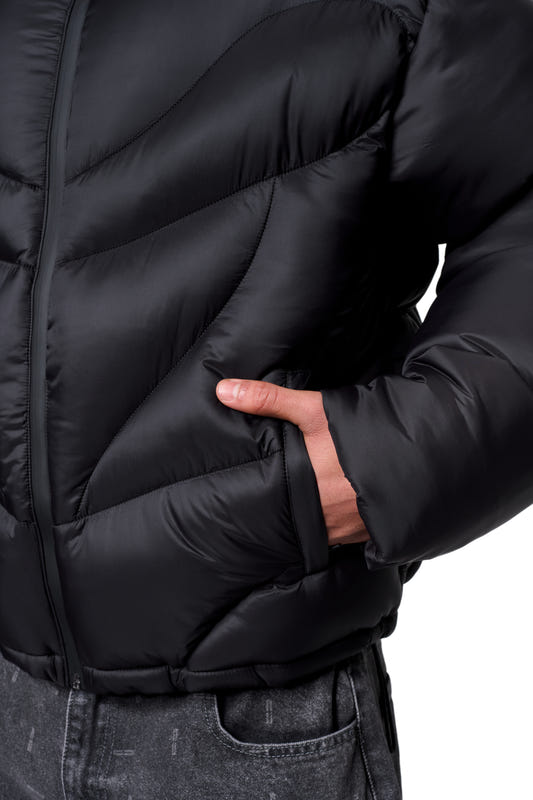A person wearing a black Void Puffer Jacket, showing the front zipper, diagonal stitching, a hand in a side pocket, and dark jeans.