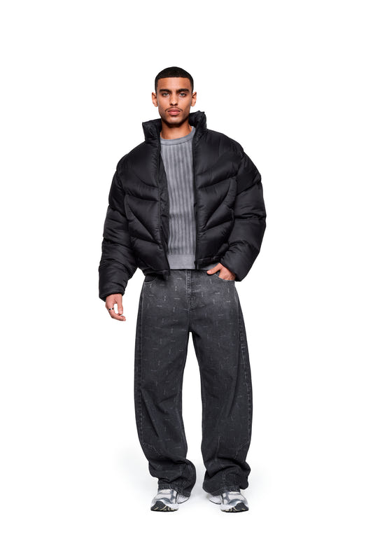 A man wears the Void Puffer Jacket open over a gray sweater, paired with loose-fitting jeans and sneakers. The jacket features a high collar and a functional design.