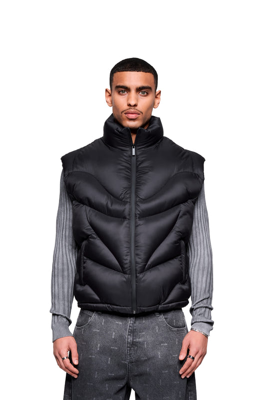 Male model wearing the Void Puffer Vest with a voluminous silhouette over a gray sweater and jeans. Showcases the fit and design of the sleeveless vest.