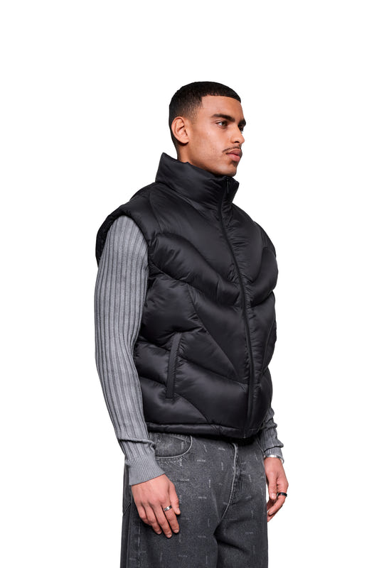 Male model wearing the Void Puffer Vest, a sleeveless, voluminous silhouette with a zipper. Paired with a gray sweater and jeans, accessorized with a silver ring and a watch.