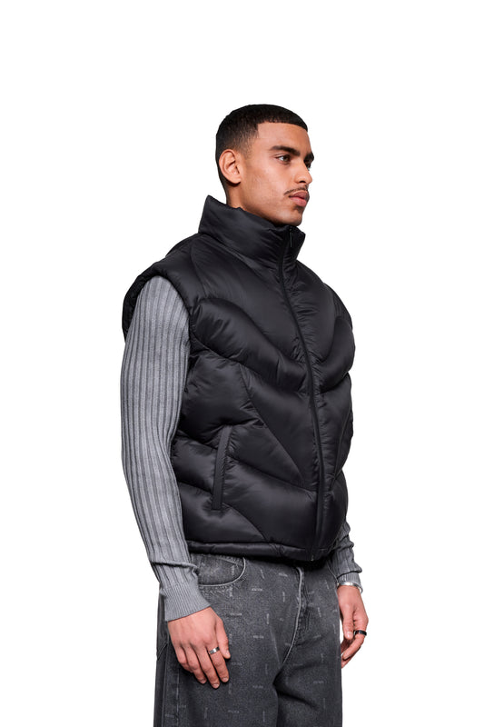Männliches Modell trägt Void Puffer Vest, ärmellose, voluminöse Silhouette mit Reißverschluss. Kombiniert mit grauem Pullover und Jeans, Silberring und Uhr als Accessoires.