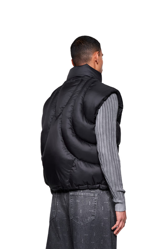 A man wears a Void Puffer Vest with contoured lines and a drawstring over a gray top and jeans, accentuated by its voluminous, sleeveless silhouette.
