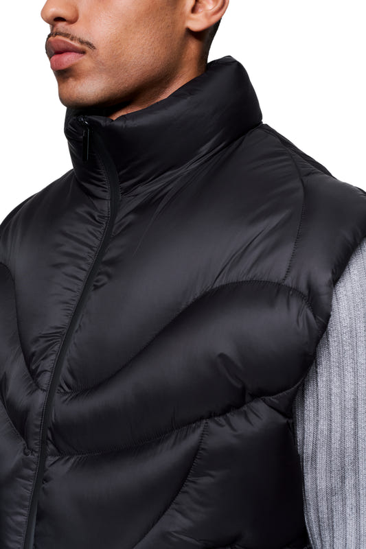 Close-up of the Void Puffer Vest, a sleeveless, voluminous vest with a high collar, worn over a gray ribbed long-sleeve sweater.