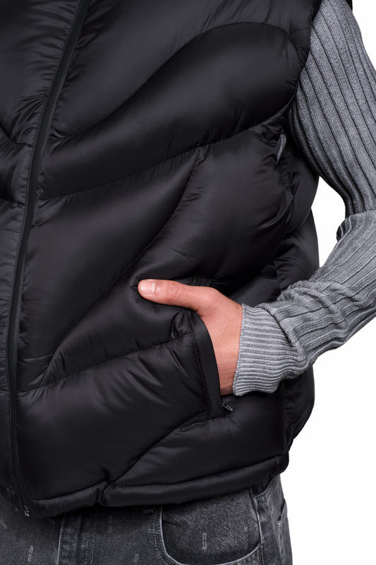 A person is wearing the Void Puffer Vest—a sleeveless, roomy quilted vest with an adjustable hem—over a gray knit sweater, with a hand in the side pocket.