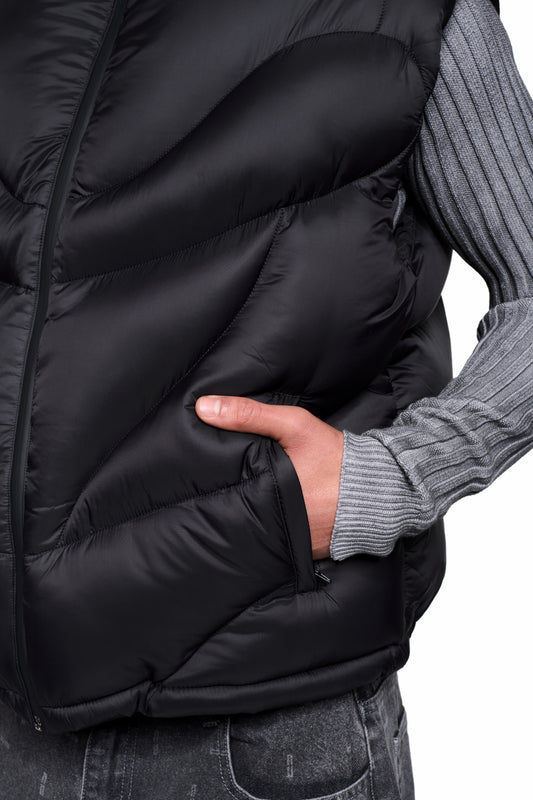 Person trägt die Void Puffer Vest, eine ärmellose, voluminöse Steppweste mit verstellbarem Saum, über einem grauen Strickpullover, Hand in der Seitentasche.