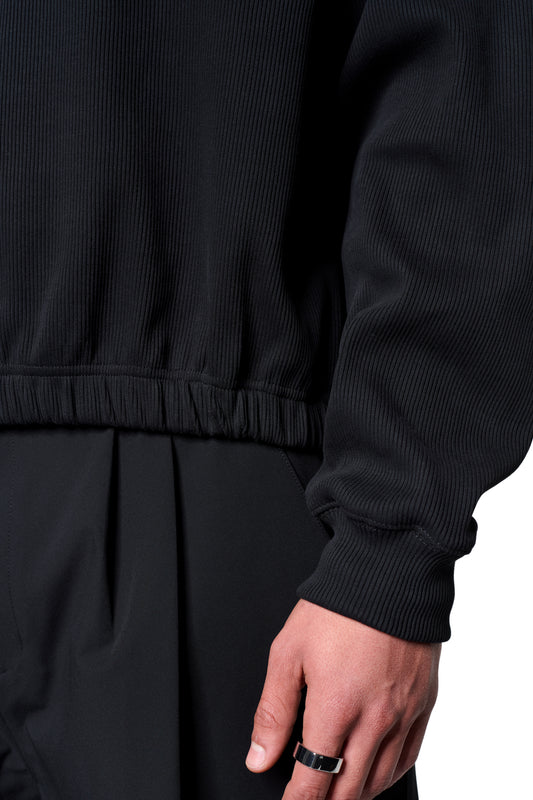 Modell trägt Void Rib Hoodie mit markanter Rippstruktur und elastischem Saum, kombiniert mit schwarzen Hosen. Rechter Mittelfinger trägt schlichten silbernen Ring.