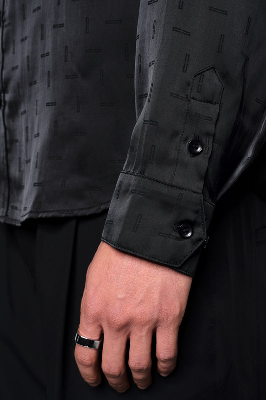 Nahaufnahme eines Arms in einem Satin Shirt Black mit eingewebtem Monogramm und doppeltem Kragen. Sichtbar sind auch eine silberne Ring und schwarze Hosen.