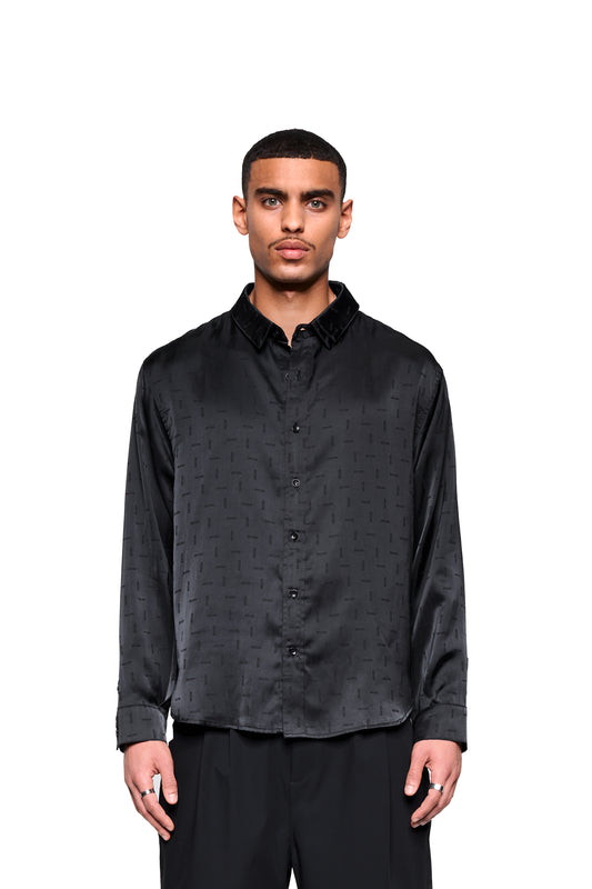 Mann trägt ein unisex Satin Shirt Black mit eingewebtem Icon-Monogramm, Doppelkragen links, abgerundetem Saum. Lockerer Sitz über schwarzen Hosen.