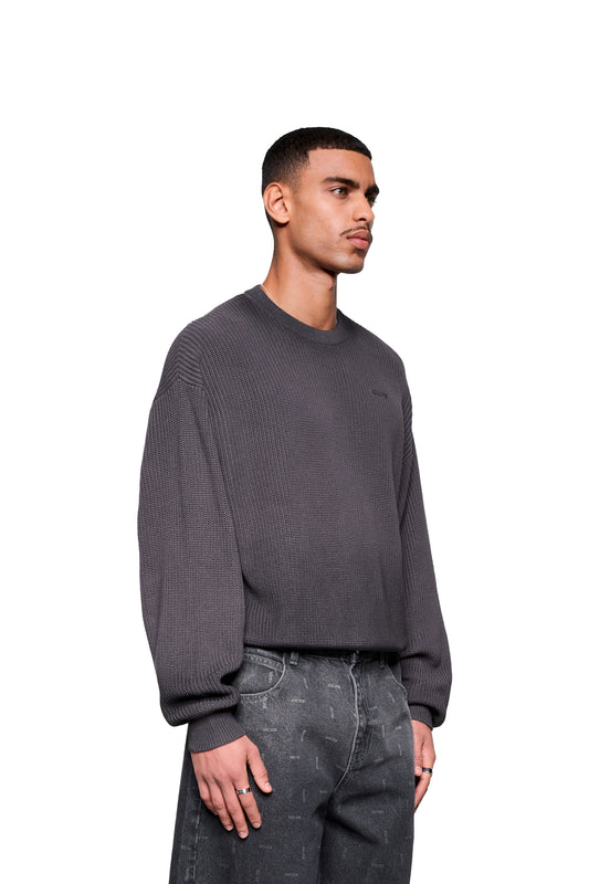 Junger Mann trägt den Void Knit Sweater Black mit markanter Struktur und cropped, boxy Form, kombiniert mit locker sitzender, dezent gemusterter Jeans.