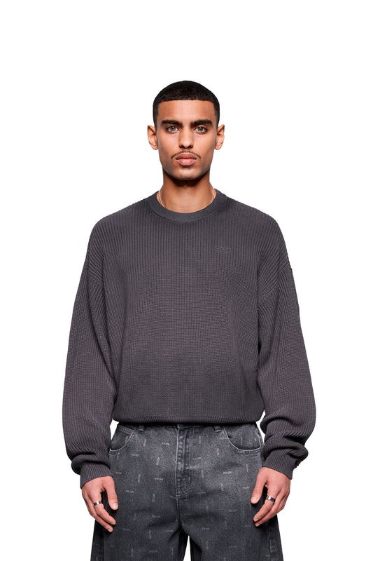 Junger Mann trägt den Void Knit Sweater Black, strukturiertes Waffelstrick-Design, moderne cropped Form und lässig geschnittene Jeans mit subtilen Prints.