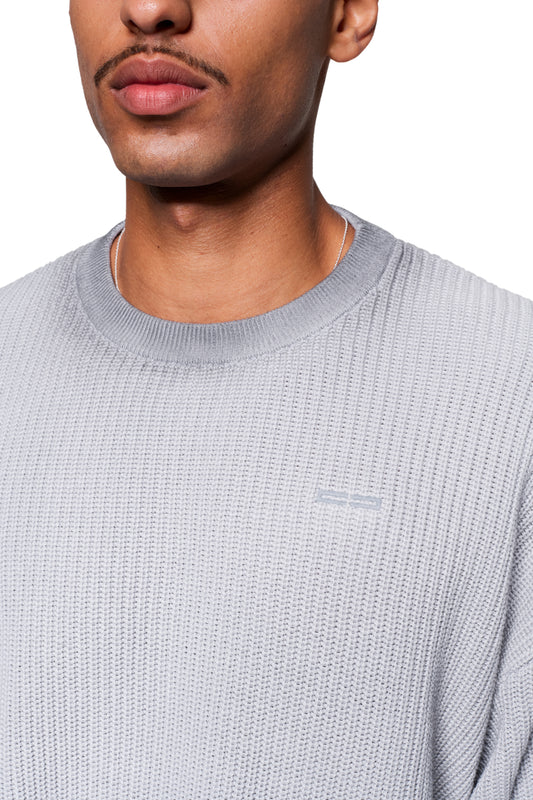 Person trägt Void Knit Sweater Gray mit markanter Waffelstruktur. Der Ausschnitt zeigt einen feinen Kettenanhänger. Fokus liegt auf Textur und modernem Schnitt.