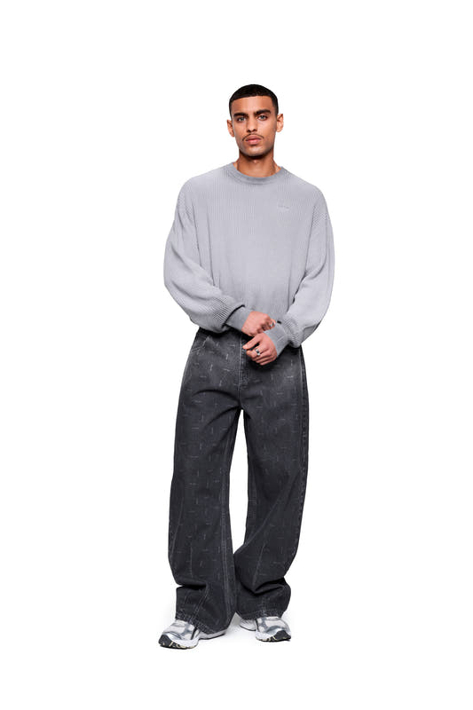 A male model wears the Void Knit Sweater in gray—cropped and boxy, with a distinctive knit texture—paired with loose-fitting black denim jeans and gray sneakers.