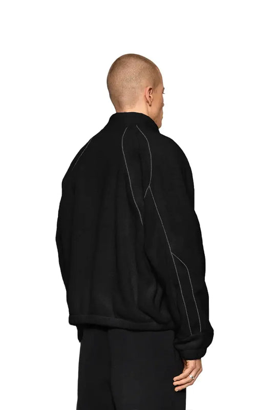 Mann trägt Reflective Fleece Jacket Black mit weißem OLA KALA Stick, reflektierenden Nähten. Jacke ist oversized, unisex, mit Gummizug an Bündchen.