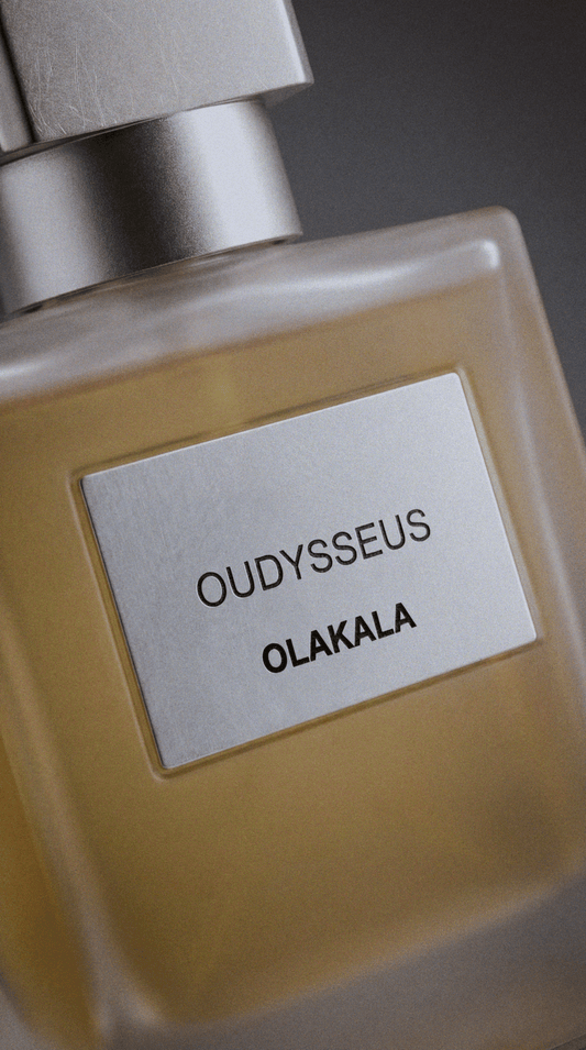Nahaufnahme einer Parfümflasche mit bernsteinfarbener Flüssigkeit, silbernem Deckel und Etikett mit ODYSSEUS und OLAKALA.