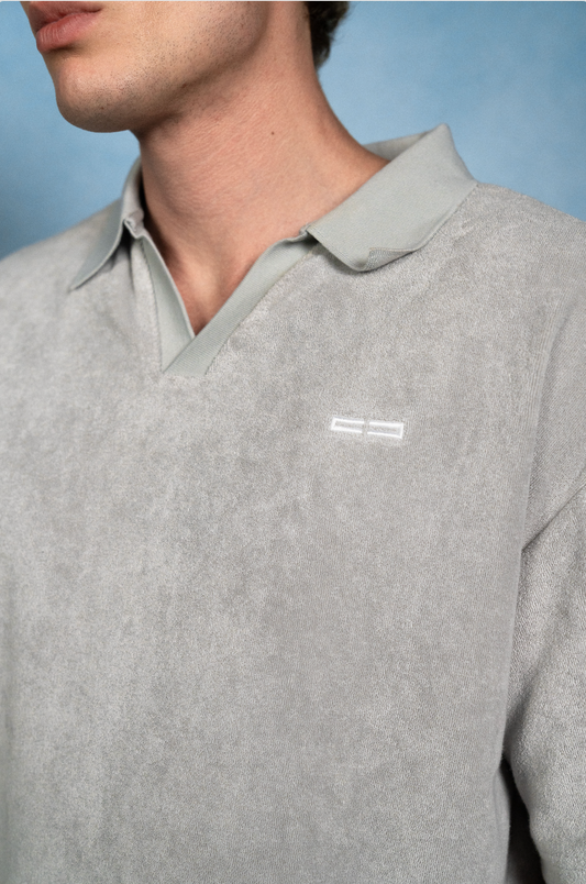 Nahaufnahme eines hellgrauen, strukturierten Polo-Pullovers mit grünem Kragen; kleines weißes Logo links auf der Brust, vor blauem Hintergrund.