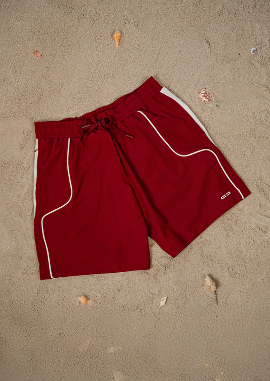 Badeshorts in Burgunderrot mit weißen Seitenpaspeln, elastischem Bund mit Kordelzug, seitliche Kontrastlinien und Logo am Bein, auf Sand liegend.