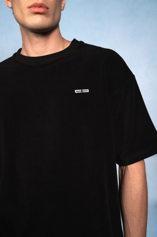 Schwarzes T-Shirt mit kleinem weißen Logo links auf der Brust, getragen von einem Mann; heller blauer Hintergrund.