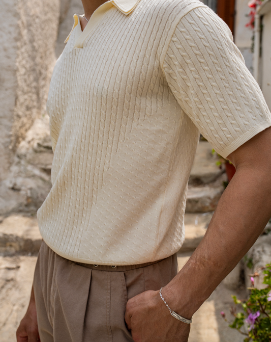 Cremefarbener, kurzärmliger Strick-Polo mit Polokragen und Zopfstrickmuster, beige Hose, silbernes Armband – Outdoor-Szene mit Steinstufen im Hintergrund.