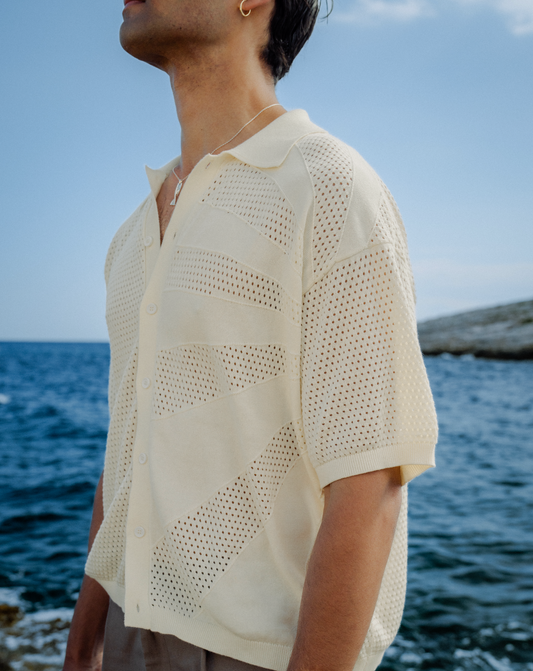 Cremefarbener Strick-Cardigan mit kurzen Ärmeln, durchbrochenem Muster und Knopfleiste, am Strand vor dem Meer.