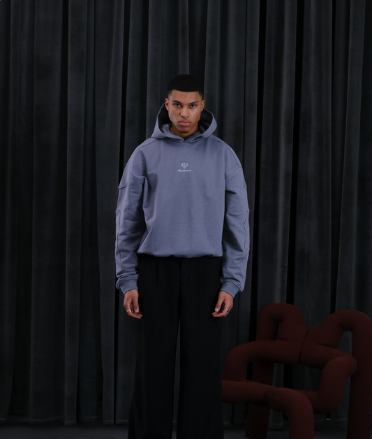 Mann in grauem Hoodie mit Logo, schwarze Hose, posiert vor dunklem Vorhang – Produktaufnahme.