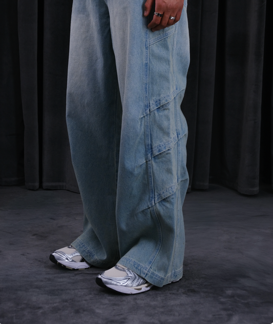 Weite hellblaue Jeans mit Panel- und Cargo-Nähten am Bein, getragen mit Sneakers; dunkler Hintergrund.