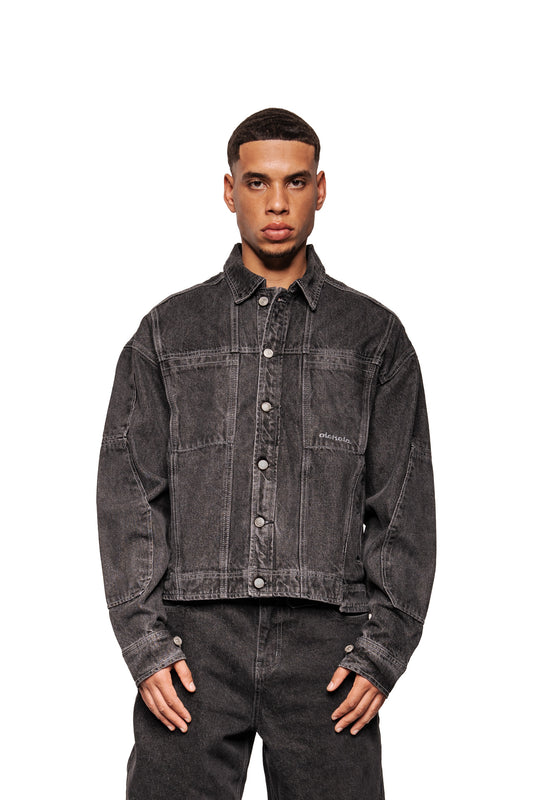 Ein männliches Modell trägt die CC Black Denim Jacket mit überschrittener Passform, klassischen Kragen, silbernen Knöpfen und gesticktem OLAKALA Logo auf der linken Brusttasche.