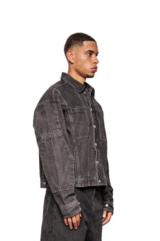 Männliches Modell im Profil mit CC Black Denim Jacket, oversized mit Panelnähten, silbernen Knöpfen, Brusttaschen und gesticktem OLAKALA Logo.