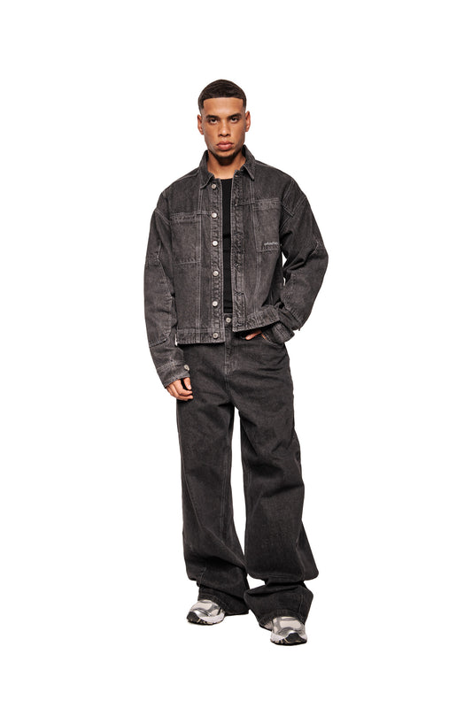 Männliches Modell in OLAKALA CC Black Denim Jacket mit Knöpfen, Brusttaschen, gesticktem Logo und locker geschnittenen Jeans. Sneaker ergänzen den lässigen Look.