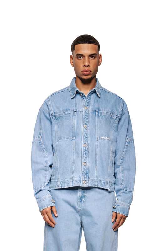 Modell trägt CC Blue Denim Jacket: lockere Passform, Knopfleiste, klassische Teilungsnähte, verstellbare Manschetten, Brust- und Vordertaschen, gesticktes OLAKALA-Logo.