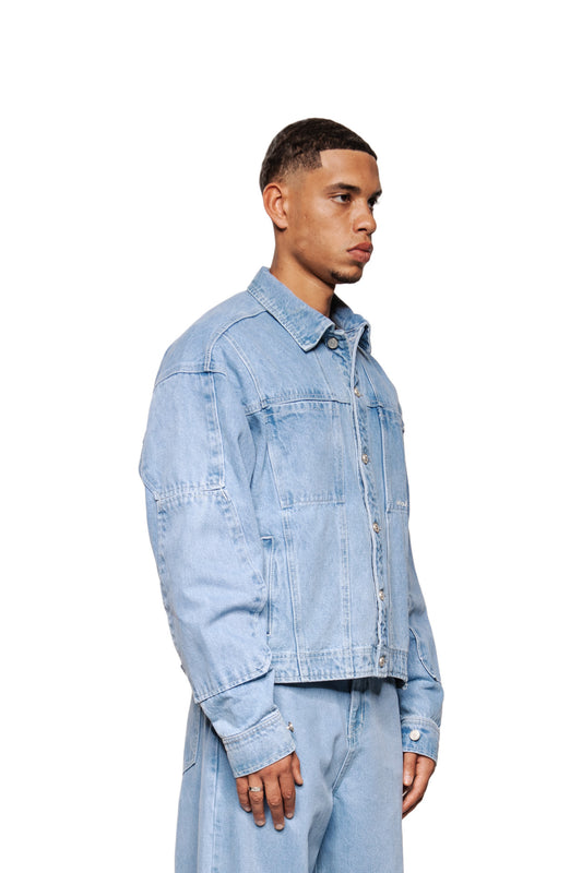 Männliches Model trägt die CC Blue Denim Jacket von OLAKALA mit silbernen Knöpfen, Brusttaschen, gesticktem Logo und verstellbarem Bund, kombiniert mit passenden Jeanshosen.