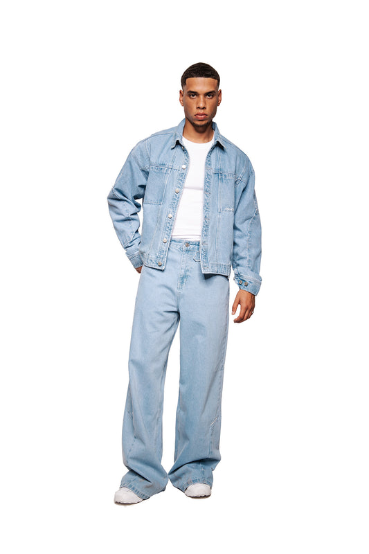 Young man in OLAKALA CC Blue Denim Jacket, loose cut with breast pockets and embroidered logo, combined with wide cut jeans and white sneakers.