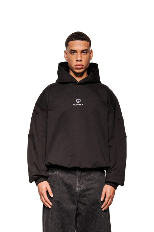 Männliches Modell trägt das Discreet Mask Hoodie Black mit OLAKALA-Logo, Rippbündchen und elastischem Saum. Kombiniert mit geraden, dunkel gewaschenen Jeans.