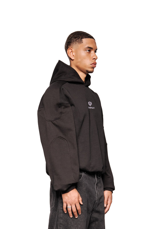 Männliches Modell trägt Discreet Mask Hoodie Black mit gesticktem Defect Identity-Icon, Panel-Details an Ellbogen/Schultern, Oversized-Schnitt und elastischen Bündchen, in Seitenansicht.