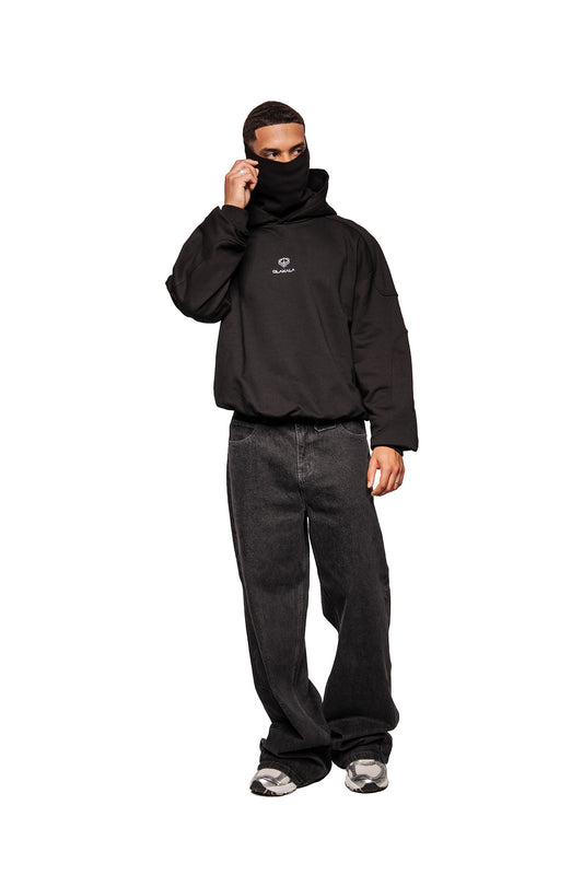 Male model wears a Discreet Mask Hoodie Black with integrated face covering, embroidered Defect Identity Icon and oversized cropped fit, combined with baggy jeans and sneakers.