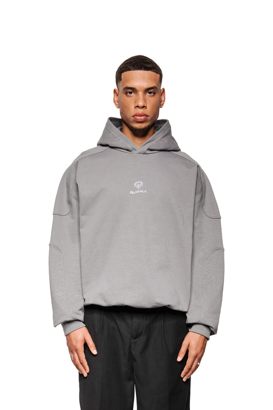 Modell trägt Discreet Mask Hoodie Dunkelgrau mit gesticktem OLAKALA-Logo, Oversize-Schnitt, Rippbündchen und Teilungsnähten, kombiniert mit geraden schwarzen Hosen.