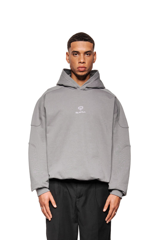 Modell trägt Discreet Mask Hoodie Dunkelgrau mit gesticktem OLAKALA-Logo, Oversize-Schnitt, Rippbündchen und Teilungsnähten, kombiniert mit geraden schwarzen Hosen.
