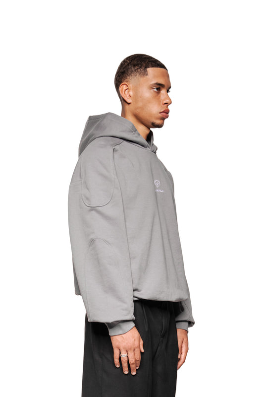 Junger Mann im Profil trägt den Discreet Mask Hoodie Dark Gray mit gesticktem OLAKALA Icon, Ripp-Bündchen und Teilungsnähten, kombiniert mit locker sitzenden schwarzen Hosen.