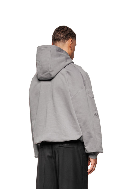 Person von hinten in einem oversized, dunkelgrauen Discreet Mask Hoodie mit Teilungsnähten und gesticktem OLAKALA-Icon, trägt lockere schwarze Hosen, gegen weißen Hintergrund.