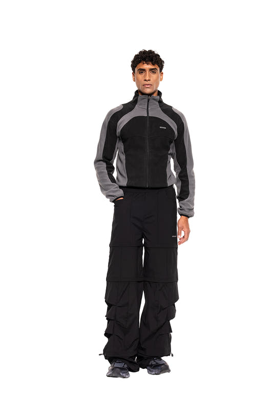 Person trägt die Light Fleece Jacket Black & Gray mit ergonomischen Flatlock-Nähten, kombiniert mit locker sitzenden, mehrlagigen schwarzen Hosen und dunklen, klobigen Sneakers.