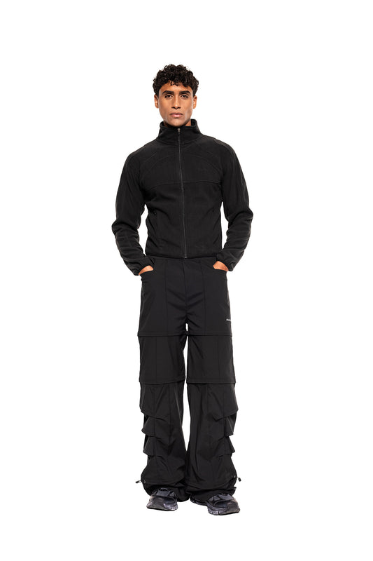 Männliches Modell trägt die Light Fleece Jacket Black von OLAKALA, kombiniert mit einzigartigen Cargo-Hosen und klobigen Sneakers, ideal als atmungsaktive Midlayer für kalte Tage.