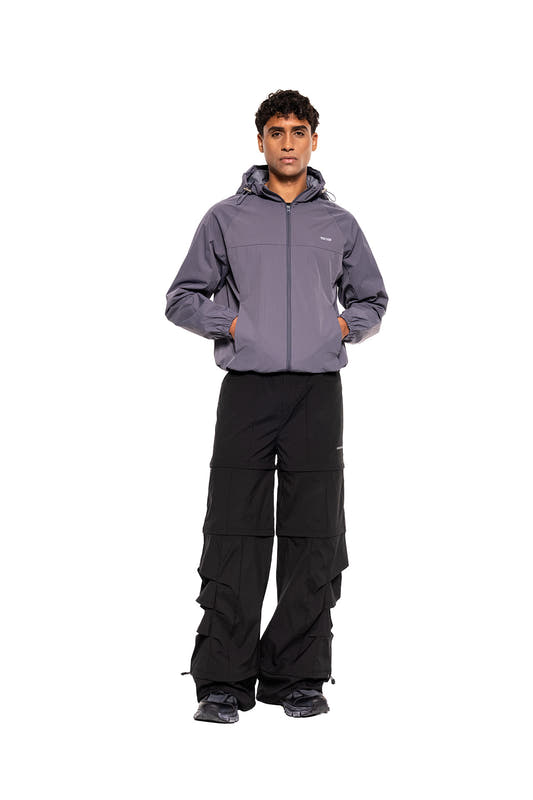 Junger Mann im OLAKALA Windbreaker Icon Gray, sportlich, funktional, mit Kapuze und Saumkordelzügen, kombiniert mit schwarzen Hosen und dunklen Sneakers für urbanen Stil.