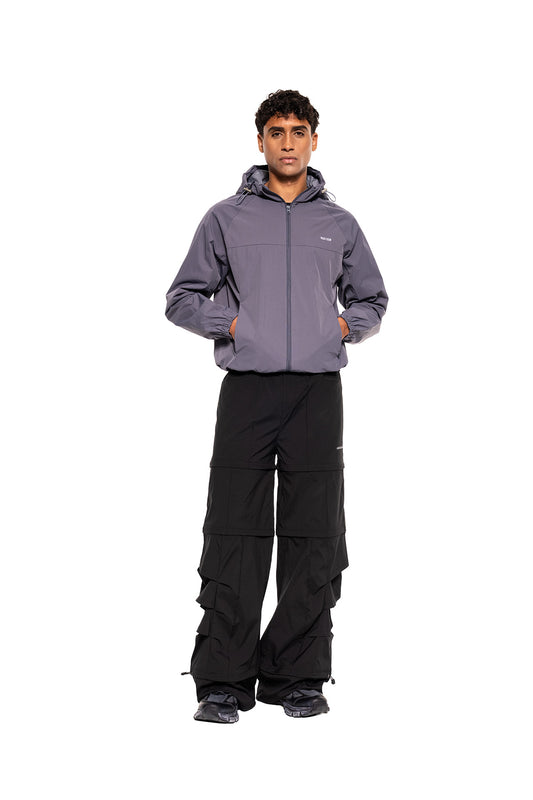 Young man in the OLAKALA Windbreaker Icon Gray, sporty, functional, with hood and hem drawstrings, combined with black pants and dark sneakers for urban style.