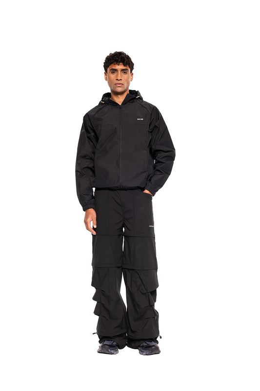 Mann trägt den Windbreaker Icon Black: sportlicher Schnitt, Kapuze mit Kordelzügen, ideal für Outdoor-Aktivitäten. Kombiniert mit cargo-inspirierten Hosen und Sneakers.