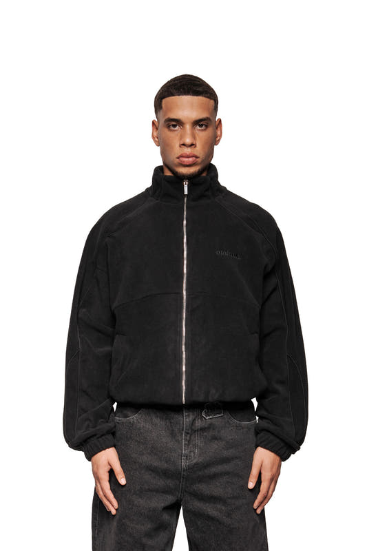Junger Mann trägt Fleece Jacket Black Onyx mit OLA KALA-Stickerei, elastischen Bündchen, paspelierten Teilungsnähten und Taschen, präsentiert im oversized Unisex-Stil.