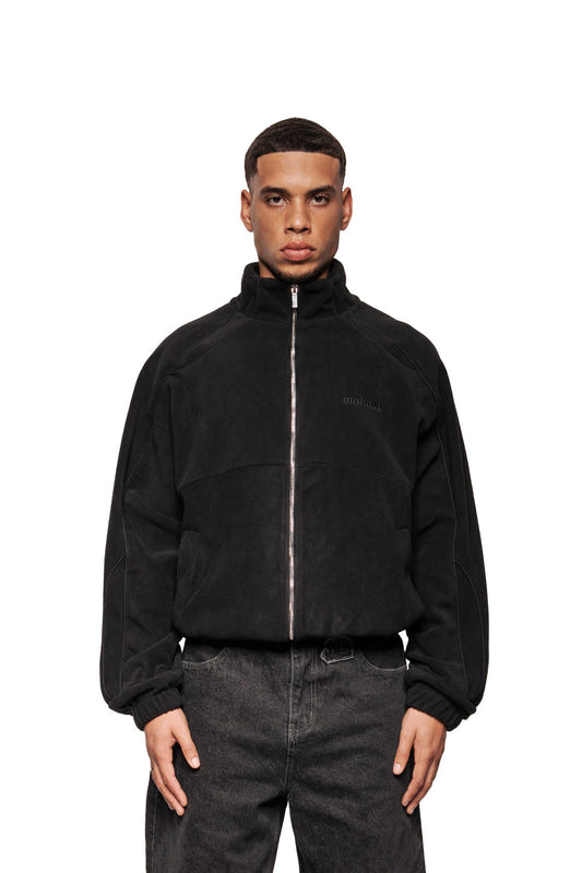 Young man wears Fleece Jacket Black Onyx with OLA KALA embroidery, elasticated cuffs, piped dividing seams and pockets, presented in an oversized unisex style.