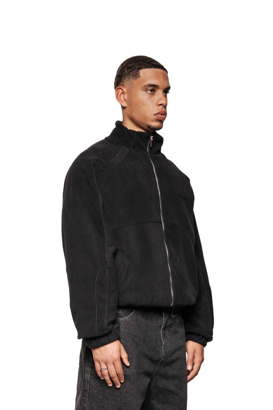 Männliches Model trägt die Fleece Jacket Black Onyx mit hohem Kragen, OLA KALA-Stickerei und Eingriffstaschen, kombiniert mit dunklen Jeans.