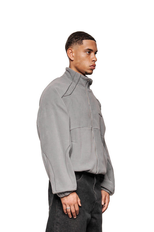 Young man in profile with fleece jacket Night Owl, gray zip-up with OLA KALA logo, dividing seams, slit pockets, and elastic cuffs, presents minimalist style.